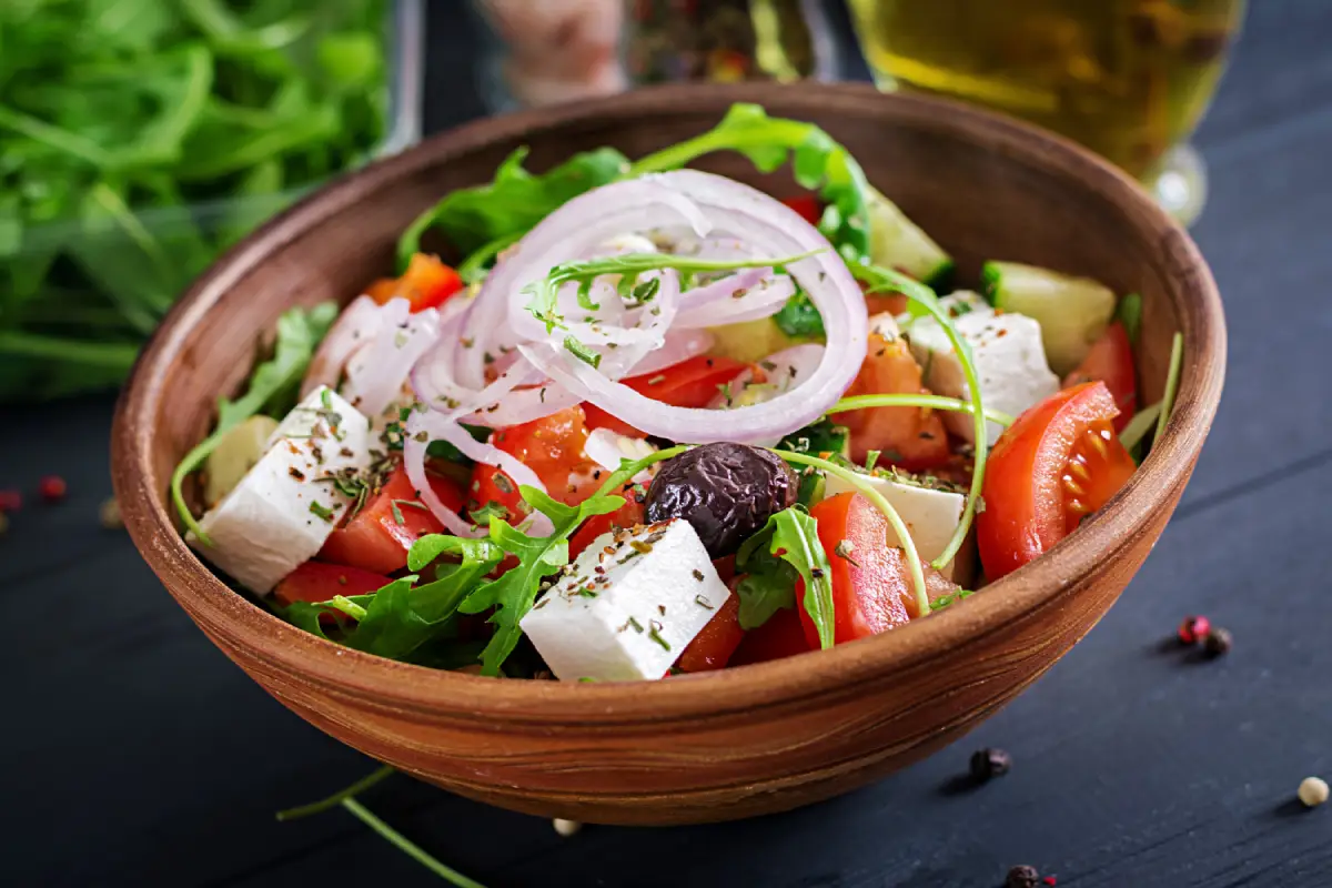 Traditional Greek salad served in a rustic earthenware bowl with ripe tomatoes, sliced onions, cucumbers, feta cheese, and olives, garnished with fresh herbs and olive oil.