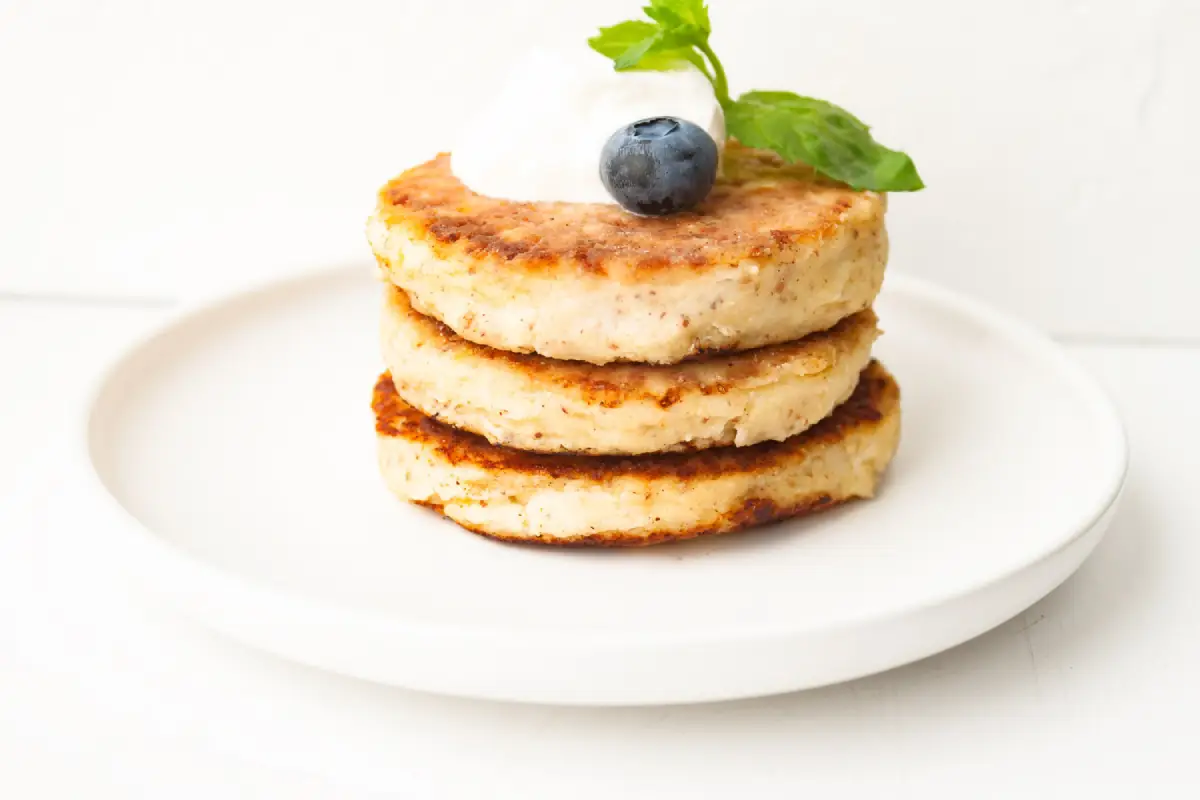 A stack of three cottage cheese pancakes with a blueberry and cream topping on a white plate.