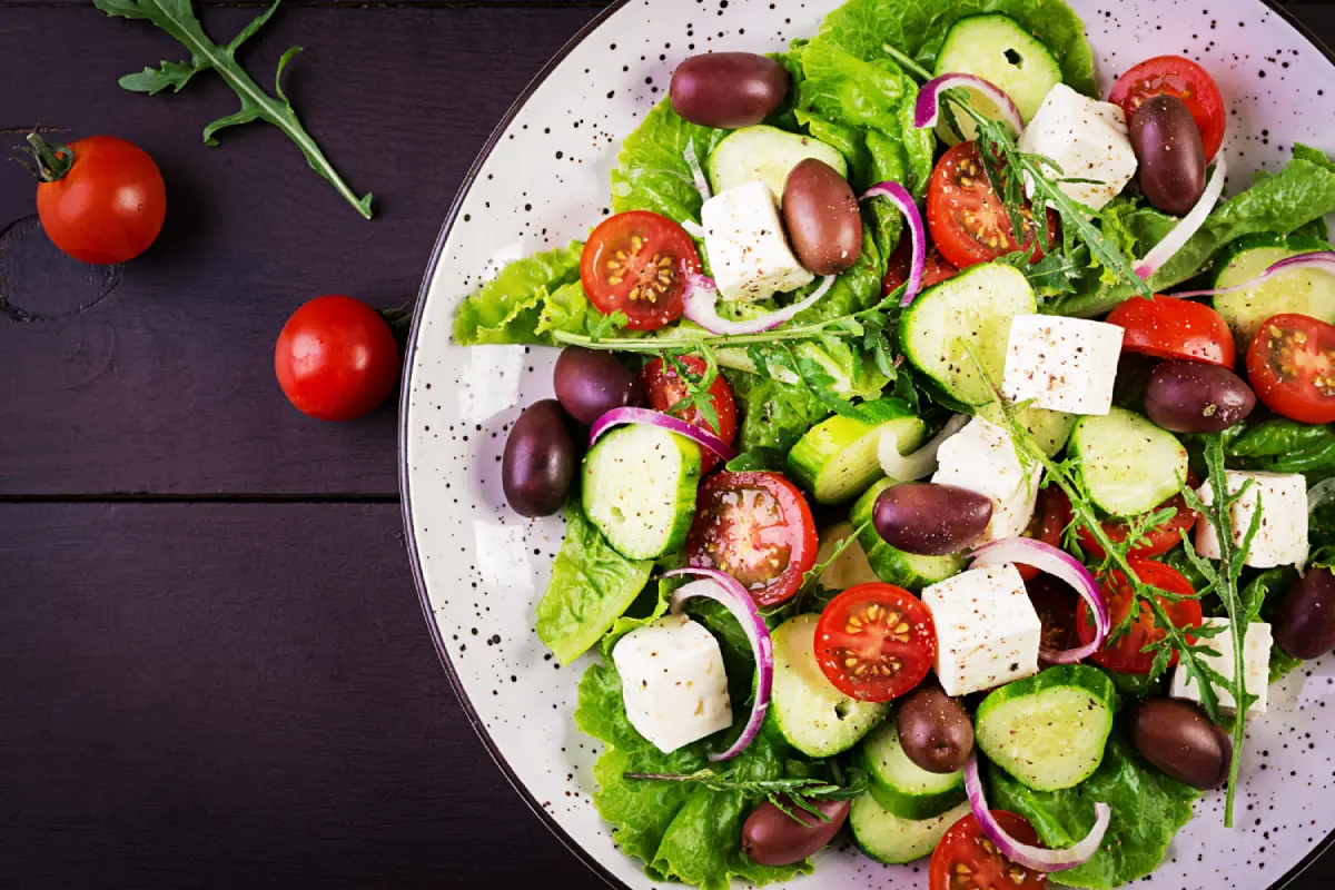 Overhead view of a classic Greek salad with crisp lettuce, juicy cherry tomatoes, cucumber slices, red onion, feta cheese, and Kalamata olives on a speckled plate.
