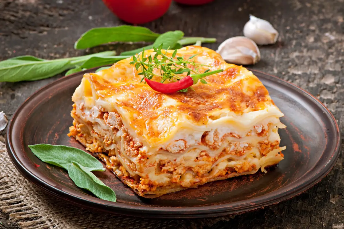 A slice of classic lasagna with bolognese sauce on a rustic plate garnished with herbs.