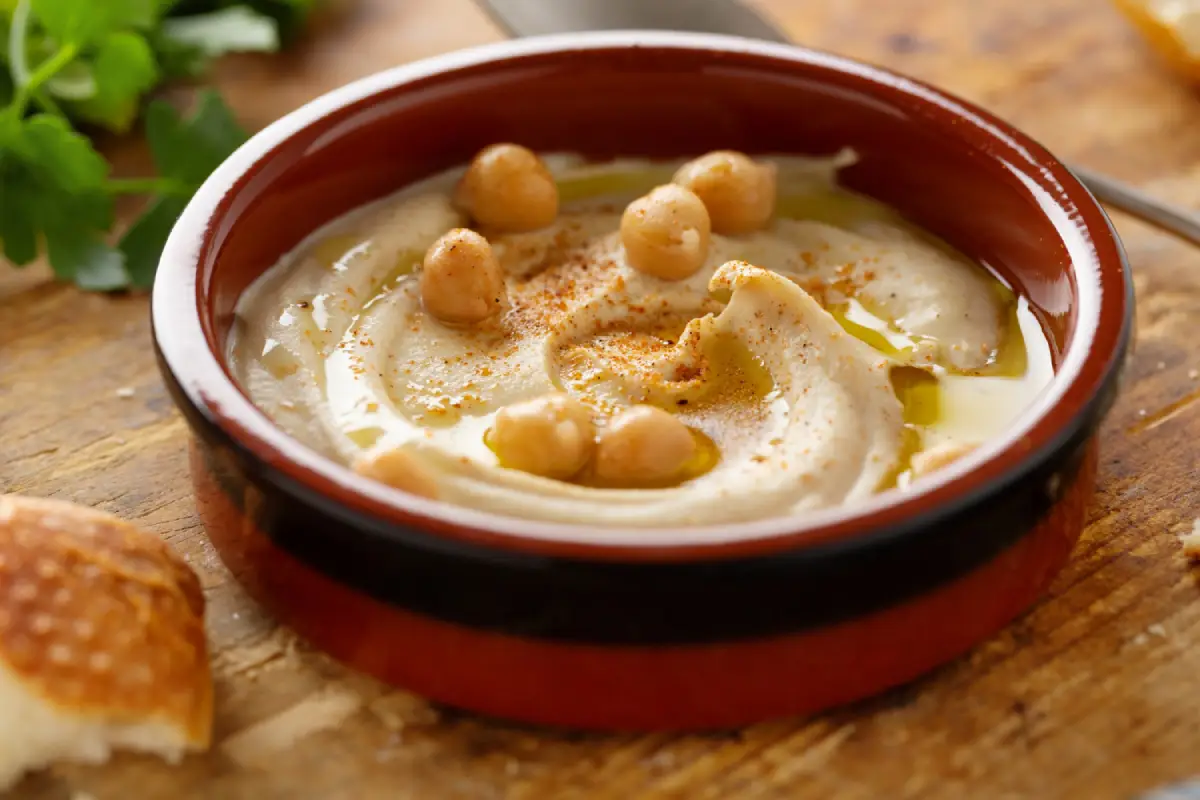 Smooth and creamy hummus in a rustic bowl, garnished with whole chickpeas, a drizzle of olive oil, and a dash of paprika, served with a piece of bread.