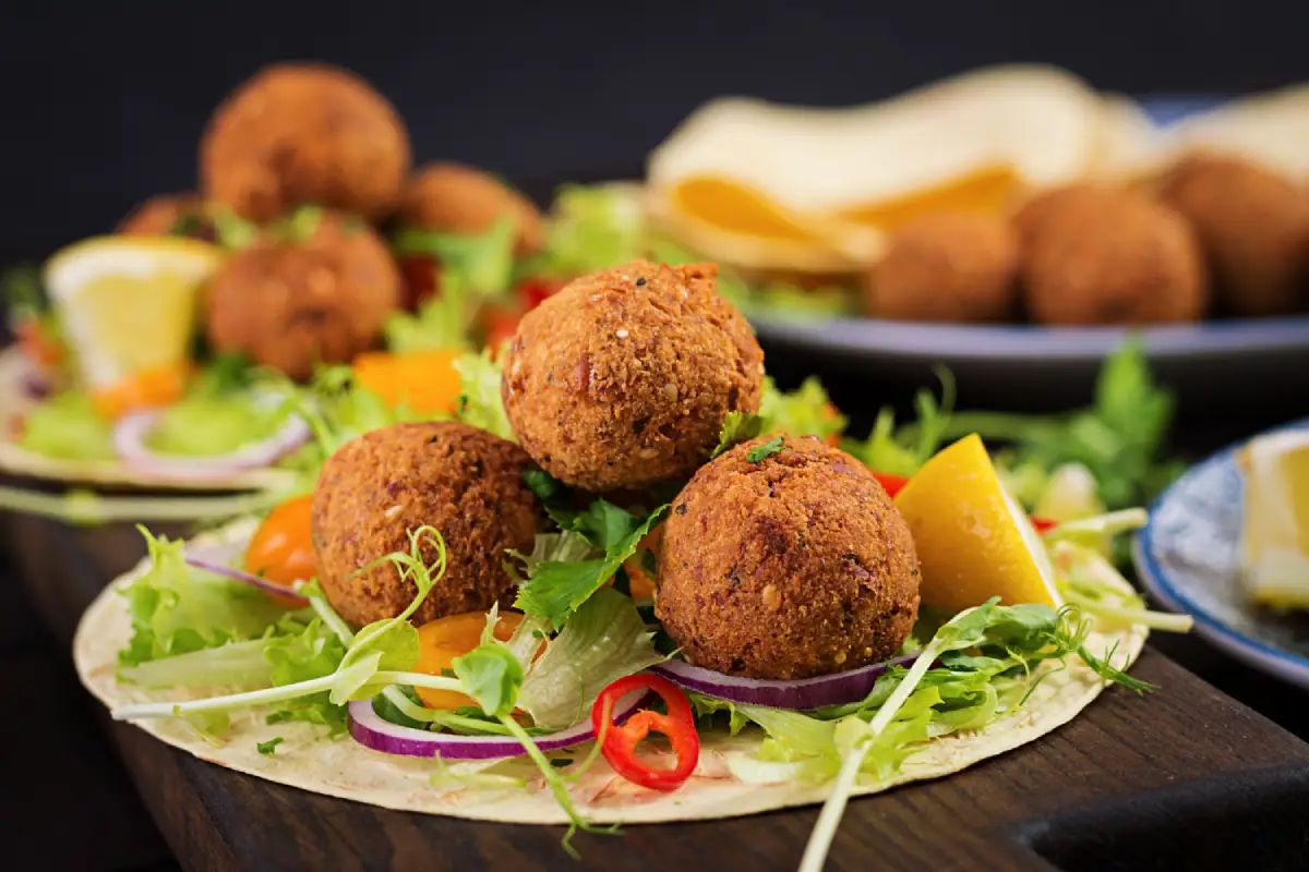 Freshly prepared falafel balls served on a bed of lettuce with lemon wedges, red onion rings, and chili slices, with soft pita bread in the background.