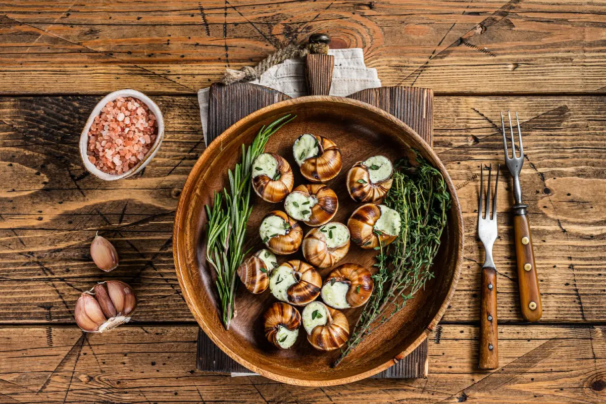 A wooden plate of French escargot garnished with garlic butter and fresh herbs on a rustic table.