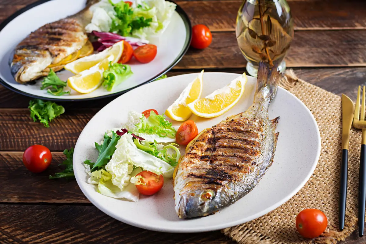 Grilled whole fish served with fresh salad and lemon slices on a rustic wooden table.