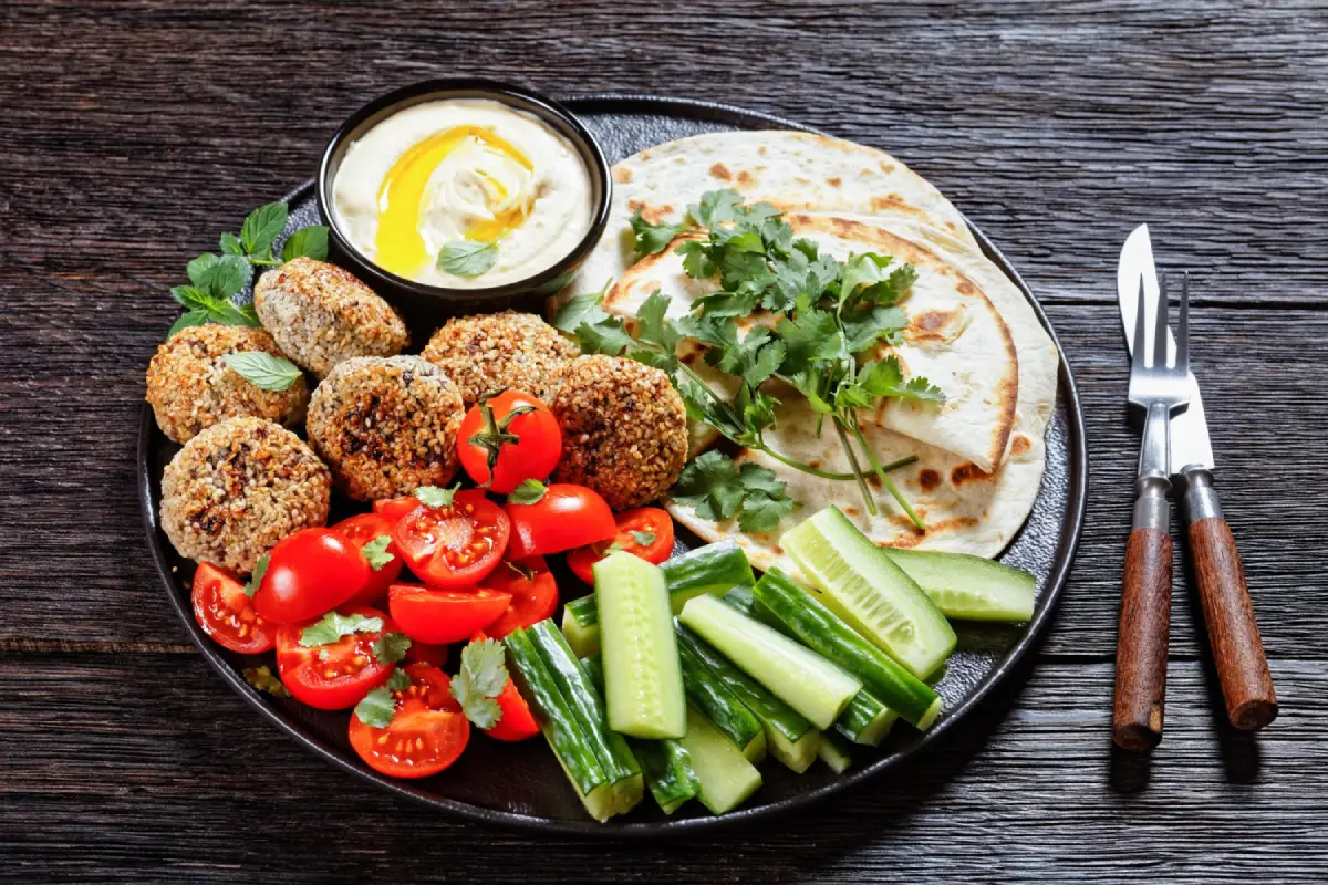 A savory platter of golden-brown falafel balls, smooth hummus with olive oil, fresh vegetables, and warm pita bread on a dark rustic table.