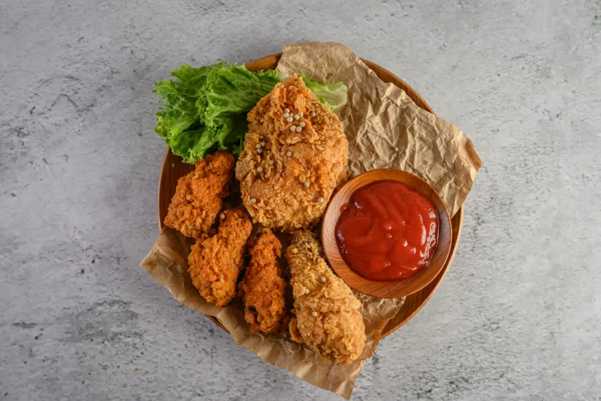 Overhead view of crispy fried chicken pieces on a wooden plate, paired with tomato sauce and a fresh green lettuce garnish, on a textured grey background.