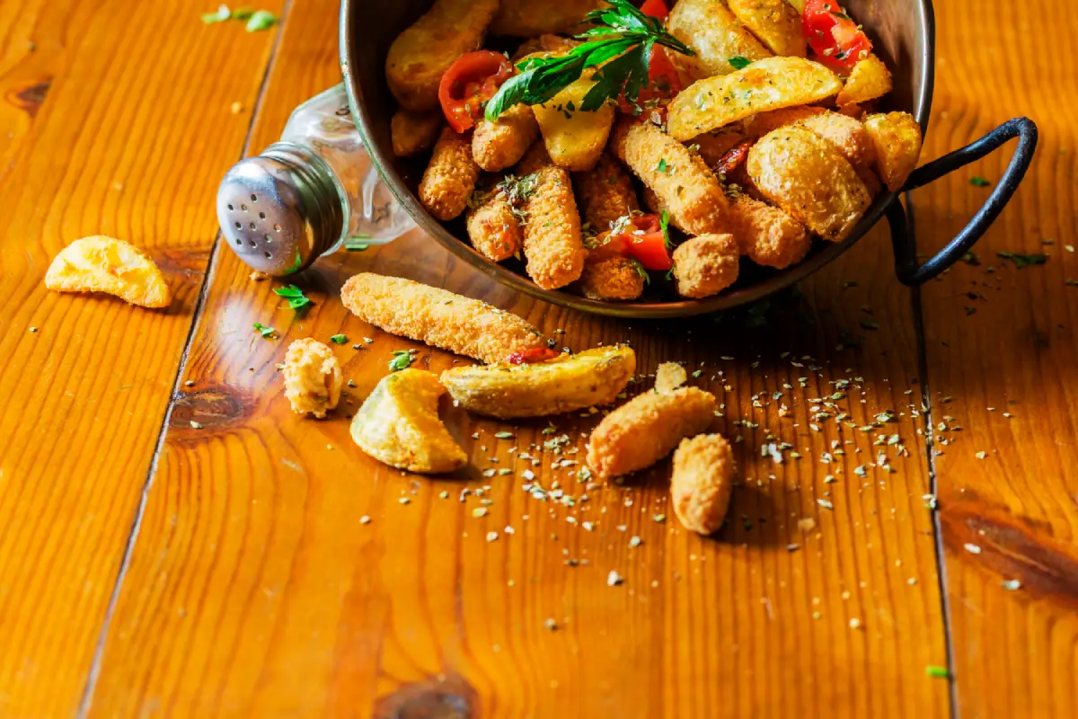 A delicious spill of golden crispy parmesan potatoes with herbs on a wooden surface, alongside an overturned salt shaker.