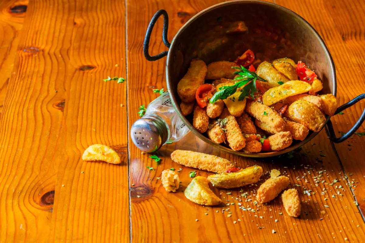 A rustic pot tipped over, spilling golden crispy parmesan potatoes with herbs and spices on a wooden table.
